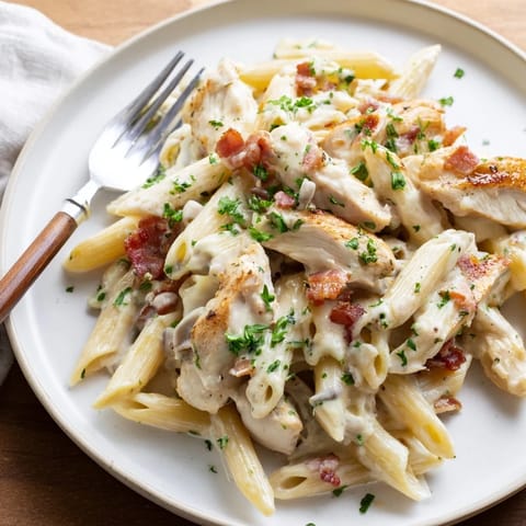 A close-up of Creamy Chicken Bacon Penne in a white bowl, highlighting the glossy garlic-Parmesan sauce coating each tube of pasta.